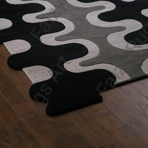 May include: A close-up of a modern area rug with a wavy pattern in black, gray, and light gray. The rug's design features flowing, organic shapes, creating a contemporary aesthetic. The rug is placed on a dark wood floor.