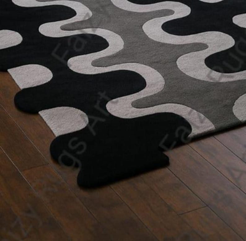 May include: A close-up of a modern area rug with a wavy pattern in black, gray, and light gray. The rug's design features flowing, organic shapes, creating a contemporary aesthetic. The rug is placed on a dark wood floor.