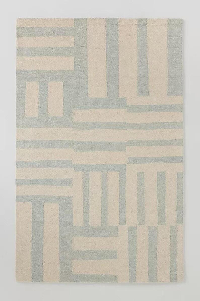 May include: Rectangular area rug with a geometric pattern. The rug features a repeating design of light blue and beige rectangular shapes. The pattern is a mix of horizontal and vertical lines, creating a modern, abstract look. The rug is made of a woven material.