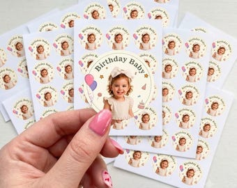 May include: A collection of circular stickers on white sheets, each featuring a photograph of a young child with a white bow, the text Birthday Baby, and illustrations of balloons, stars, and a cake. One sticker is held up to show the design detail.