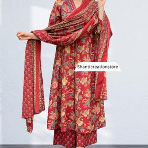 May include: A red floral print salwar kameez set. The set includes a long tunic top, wide-leg pants, and a matching scarf. The tunic has long sleeves and a V-neckline. The floral pattern features yellow, green, and pink accents. The scarf is draped over the shoulders. The text 'Shanticreationstore' is visible.
