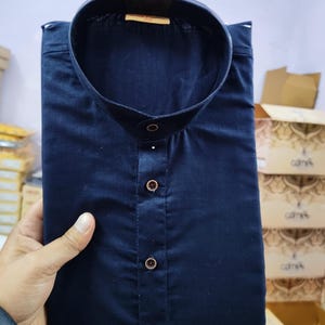 May include: A navy blue, collared shirt with a button-up front. The shirt is neatly folded. The buttons are a contrasting colour. The shirt appears to be made of a smooth fabric. The shirt is held by a hand.