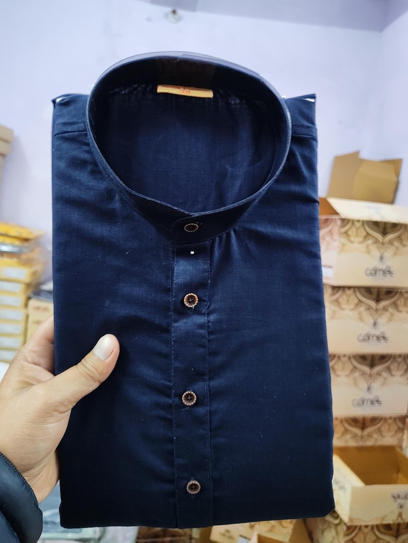 May include: A navy blue, collared shirt with a button-up front. The shirt is neatly folded. The buttons are a contrasting colour. The shirt appears to be made of a smooth fabric. The shirt is held by a hand.