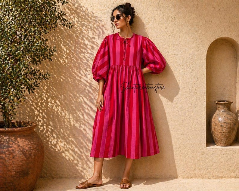 May include: A midi-length dress with vertical stripes in shades of red and pink. The dress has a round neckline with a tie, and voluminous, elbow-length sleeves. The model is wearing sunglasses and sandals.