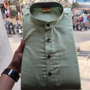 May include: A sage green shirt with a mandarin collar and three dark brown buttons. The shirt is folded neatly, with the brand name "camete" and size "38" visible on a tag. The shirt is made of a lightweight fabric.