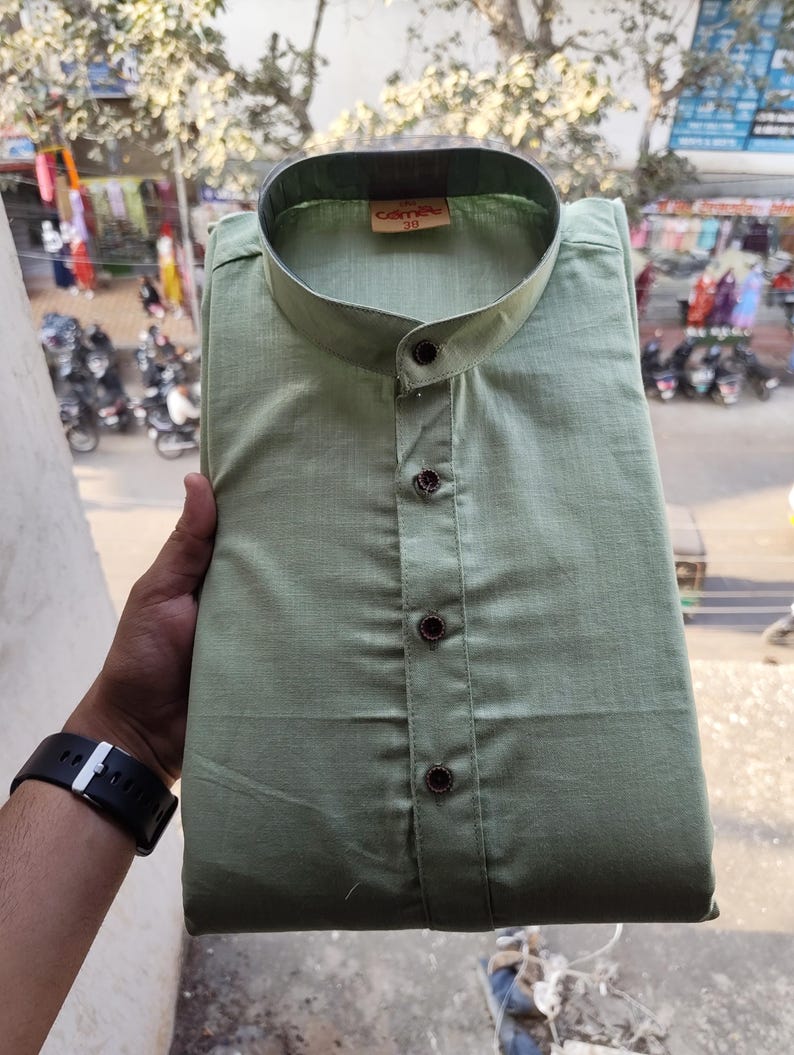May include: A sage green shirt with a mandarin collar and three dark brown buttons. The shirt is folded neatly, with the brand name "camete" and size "38" visible on a tag. The shirt is made of a lightweight fabric.