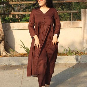 Peut inclure: Une robe marron &agrave; manches longues avec un col en V et des boutons, associ&eacute;e &agrave; un pantalon assorti. La robe pr&eacute;sente un design fluide et &agrave; plusieurs niveaux. La tenue est compl&eacute;t&eacute;e par des ballerines &agrave; motifs. Le style g&eacute;n&eacute;ral est d&eacute;contract&eacute; et confortable.