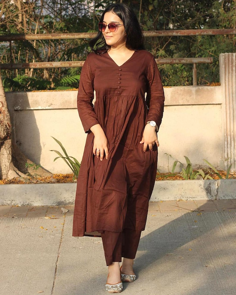 Peut inclure: Une robe marron &agrave; manches longues avec un col en V et des boutons, associ&eacute;e &agrave; un pantalon assorti. La robe pr&eacute;sente un design fluide et &agrave; plusieurs niveaux. La tenue est compl&eacute;t&eacute;e par des ballerines &agrave; motifs. Le style g&eacute;n&eacute;ral est d&eacute;contract&eacute; et confortable.