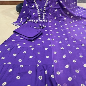 May include: A purple, long-sleeved dress with white circular patterns and a matching scarf. The dress features a lace-up neckline and a tiered skirt. The outfit is held up by a person.
