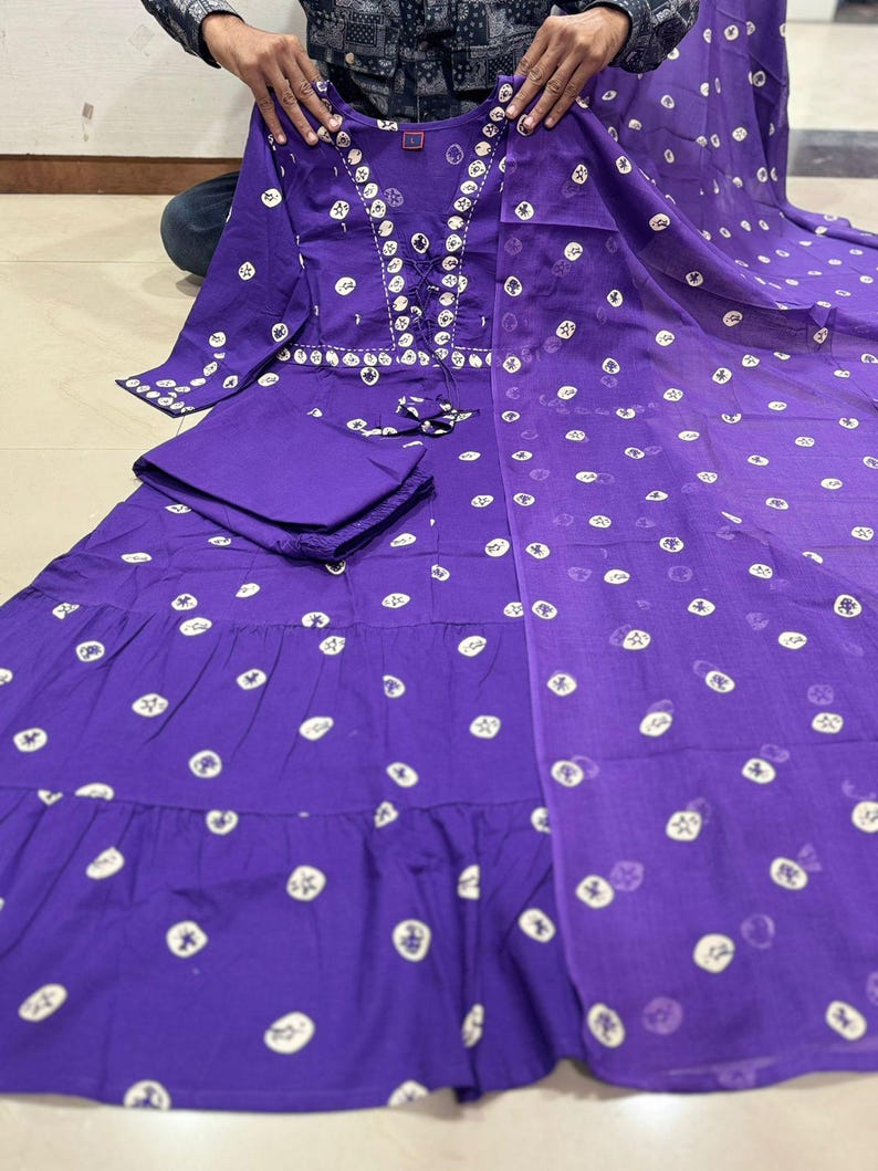 May include: A purple, long-sleeved dress with white circular patterns and a matching scarf. The dress features a lace-up neckline and a tiered skirt. The outfit is held up by a person.