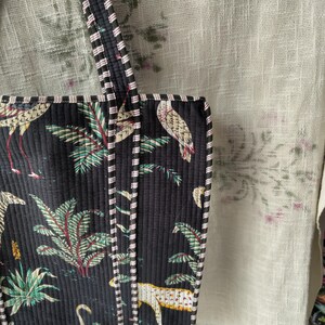 Pode incluir: Bolsa tote acolchoada preta com estampa de p&aacute;ssaros tropicais e folhagens. A bolsa apresenta acabamento e al&ccedil;as listradas. O design inclui imagens de p&aacute;ssaros, folhas de palmeira e um leopardo. A bolsa &eacute; feita de tecido acolchoado.