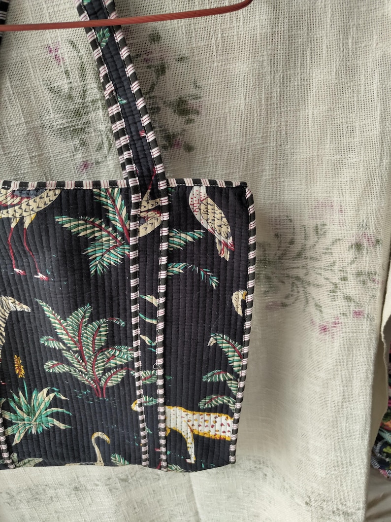 Pode incluir: Bolsa tote acolchoada preta com estampa de p&aacute;ssaros tropicais e folhagens. A bolsa apresenta acabamento e al&ccedil;as listradas. O design inclui imagens de p&aacute;ssaros, folhas de palmeira e um leopardo. A bolsa &eacute; feita de tecido acolchoado.