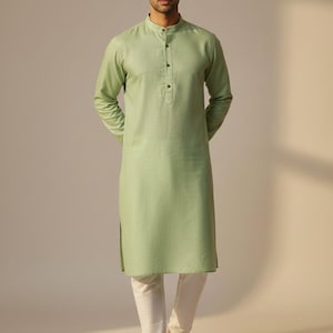 May include: A pale green kurta with a mandarin collar and long sleeves, worn with white trousers. The kurta features a button-down front and a loose fit. The outfit is finished with brown sandals.