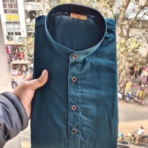 May include: Teal button-down shirt with a mandarin collar. The shirt is folded and appears to be made of a soft fabric. The buttons are a contrasting colour. The tag on the collar reads "38".