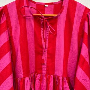 May include: A bright, striped dress in pink and red hangs on a wooden hanger. The dress has vertical stripes, a gathered waist, and a tie detail at the neckline. The sleeves are a three-quarter length.