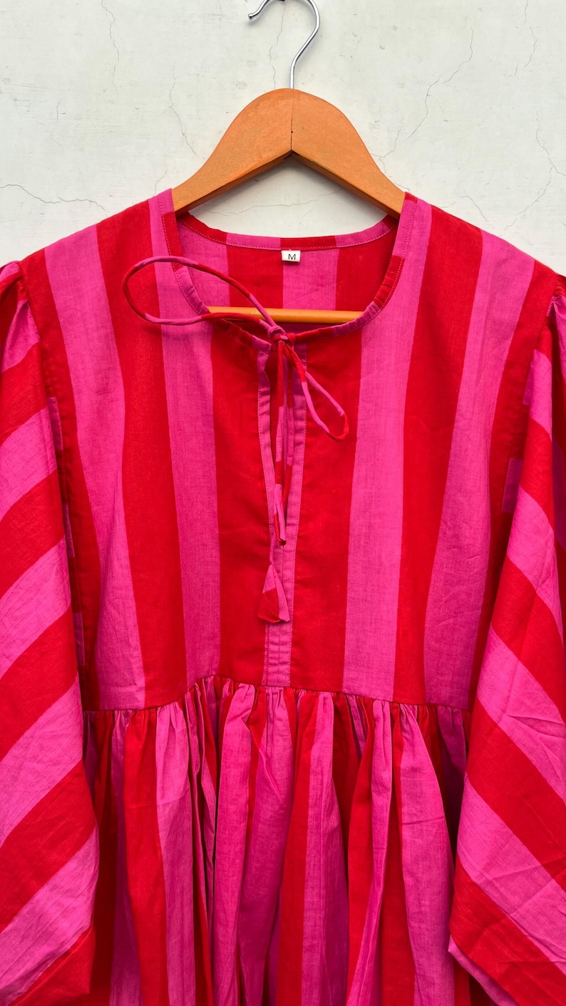 May include: A bright, striped dress in pink and red hangs on a wooden hanger. The dress has vertical stripes, a gathered waist, and a tie detail at the neckline. The sleeves are a three-quarter length.