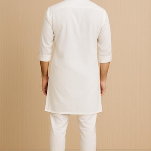 Pu&ograve; includere: Completo kurta pajama bianco. L'outfit include una lunga tunica con colletto alla coreana e maniche a tre quarti, abbinata a pantaloni dritti coordinati. Il modello indossa scarpe beige. Lo sfondo &egrave; di colore beige chiaro.