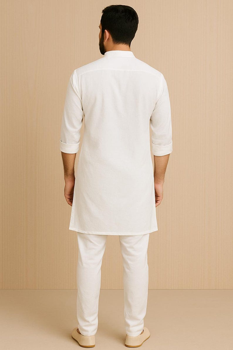 Pu&ograve; includere: Completo kurta pajama bianco. L'outfit include una lunga tunica con colletto alla coreana e maniche a tre quarti, abbinata a pantaloni dritti coordinati. Il modello indossa scarpe beige. Lo sfondo &egrave; di colore beige chiaro.