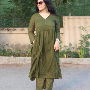 Peut inclure: Robe olive &agrave; manches longues avec un col en V et des boutons. La robe a une jupe ample et pliss&eacute;e, port&eacute;e avec un pantalon olive assorti. L'ensemble est compl&eacute;t&eacute; par des lunettes de soleil et des boucles d'oreilles argent&eacute;es.