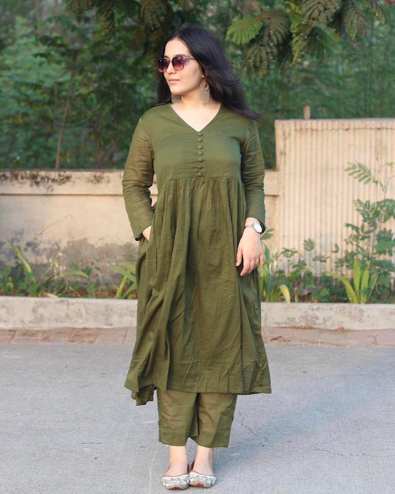 Peut inclure: Robe olive &agrave; manches longues avec un col en V et des boutons. La robe a une jupe ample et pliss&eacute;e, port&eacute;e avec un pantalon olive assorti. L'ensemble est compl&eacute;t&eacute; par des lunettes de soleil et des boucles d'oreilles argent&eacute;es.