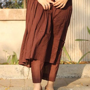 Peut inclure: Une robe pliss&eacute;e marron et un pantalon assorti sont pr&eacute;sent&eacute;s. La tenue est associ&eacute;e &agrave; des ballerines brod&eacute;es. La personne porte une montre argent&eacute;e et une bague. L'ensemble est en tissu l&eacute;ger.