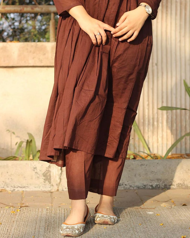 Peut inclure: Une robe pliss&eacute;e marron et un pantalon assorti sont pr&eacute;sent&eacute;s. La tenue est associ&eacute;e &agrave; des ballerines brod&eacute;es. La personne porte une montre argent&eacute;e et une bague. L'ensemble est en tissu l&eacute;ger.