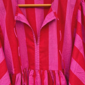 May include: A red and pink striped dress with a collared neckline and a gathered skirt. The dress is hanging on a wooden hanger. The fabric has vertical stripes in alternating shades of red and pink.