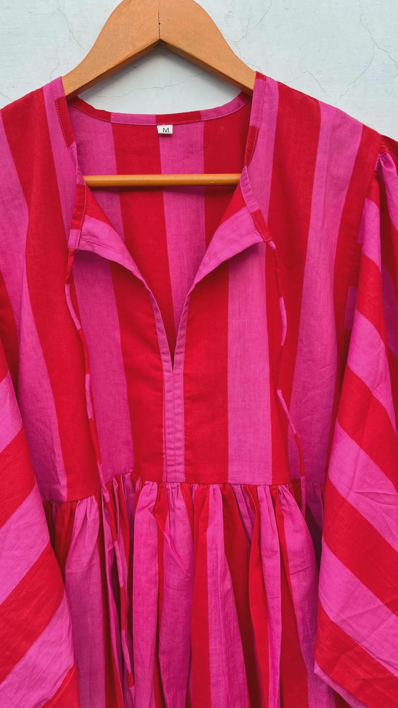 May include: A red and pink striped dress with a collared neckline and a gathered skirt. The dress is hanging on a wooden hanger. The fabric has vertical stripes in alternating shades of red and pink.