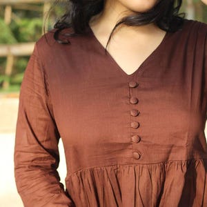 Peut inclure: Une robe marron &agrave; col en V avec des manches longues et une jupe fronc&eacute;e. La robe comporte une s&eacute;rie de petits boutons ronds sur le devant. Le tissu semble &ecirc;tre l&eacute;ger.