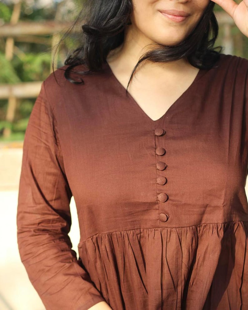 Peut inclure: Une robe marron &agrave; col en V avec des manches longues et une jupe fronc&eacute;e. La robe comporte une s&eacute;rie de petits boutons ronds sur le devant. Le tissu semble &ecirc;tre l&eacute;ger.