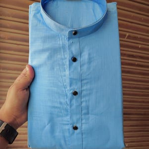 May include: A pale blue shirt with a mandarin collar and three dark buttons. The shirt is neatly folded, displaying the collar and buttoned front. The fabric looks to be a lightweight cotton or linen, suitable for warmer weather.