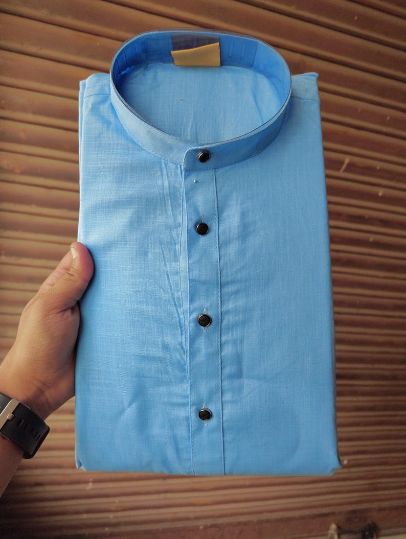 May include: A pale blue shirt with a mandarin collar and three dark buttons. The shirt is neatly folded, displaying the collar and buttoned front. The fabric looks to be a lightweight cotton or linen, suitable for warmer weather.
