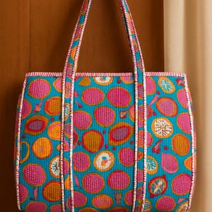 以下が含まれることがあります： ターコイズブルーの背景に、ピンク、オレンジ、紫の円が描かれたカラフルなトートバッグ。丈夫なハンドルと白いパイピングが施されています。キルティング生地で作られたバッグです。