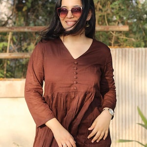 Peut inclure: Une robe marron &agrave; manches longues avec un col en V et des boutons. La robe a une taille fronc&eacute;e et une jupe fluide. La personne sur l'image porte des lunettes de soleil et une montre.