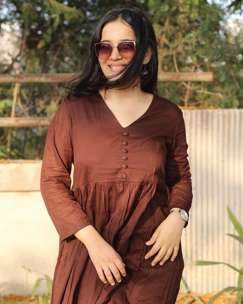Peut inclure: Une robe marron &agrave; manches longues avec un col en V et des boutons. La robe a une taille fronc&eacute;e et une jupe fluide. La personne sur l'image porte des lunettes de soleil et une montre.