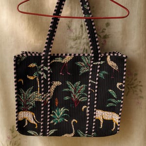 Pode incluir: Bolsa tote preta com estampa tropical com palmeiras, leopardos e p&aacute;ssaros. A bolsa tem um design acolchoado com acabamentos e al&ccedil;as listradas em preto e rosa. A bolsa est&aacute; pendurada em um cabide vermelho.