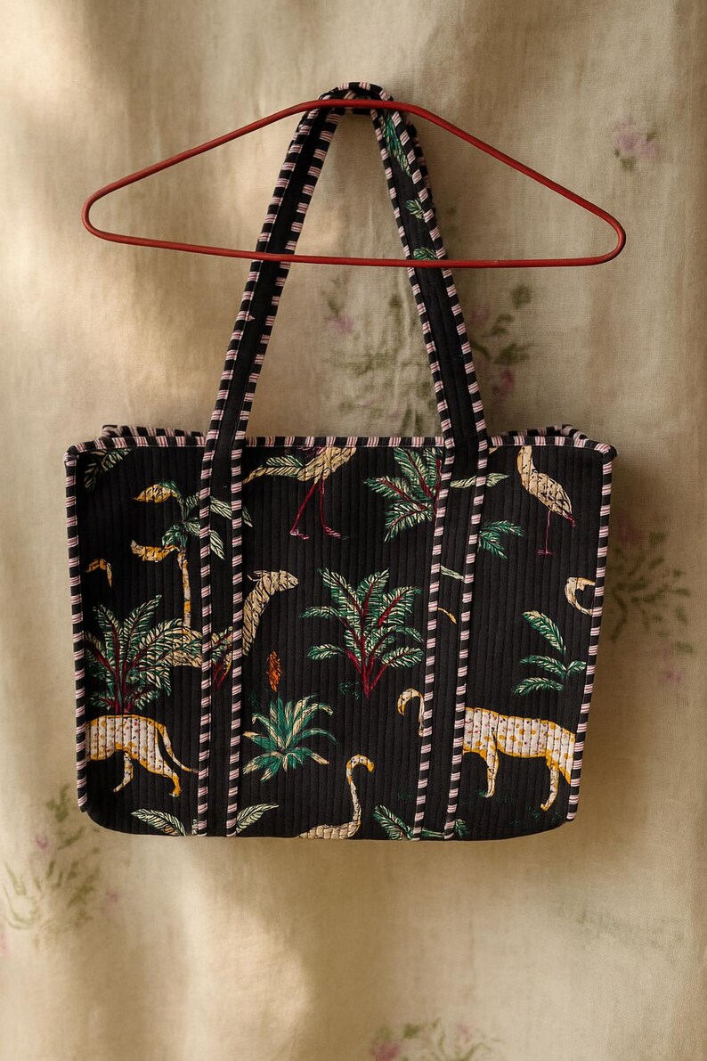 Pode incluir: Bolsa tote preta com estampa tropical com palmeiras, leopardos e p&aacute;ssaros. A bolsa tem um design acolchoado com acabamentos e al&ccedil;as listradas em preto e rosa. A bolsa est&aacute; pendurada em um cabide vermelho.