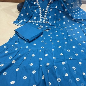 May include: A teal-colored, long-sleeved dress with a matching folded fabric square. The dress features a white, circular pattern and decorative embellishments around the neckline. The dress has a tiered skirt and is displayed on a surface.