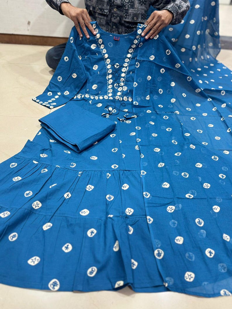 May include: A teal-colored, long-sleeved dress with a matching folded fabric square. The dress features a white, circular pattern and decorative embellishments around the neckline. The dress has a tiered skirt and is displayed on a surface.
