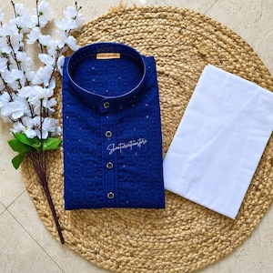 May include: A navy blue embroidered kurta shirt with a mandarin collar and button-down front, alongside white trousers. The shirt has delicate embellishments and the text "Shantireationsstore". The outfit is displayed on a woven mat with white floral accents.
