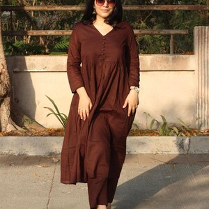 Peut inclure: Une robe marron &agrave; manches longues avec un col en V et des boutons. La robe a une jupe &eacute;vas&eacute;e et est associ&eacute;e &agrave; un pantalon assorti. La personne porte des lunettes de soleil et une montre. La tenue est adapt&eacute;e aux tenues d&eacute;contract&eacute;es.