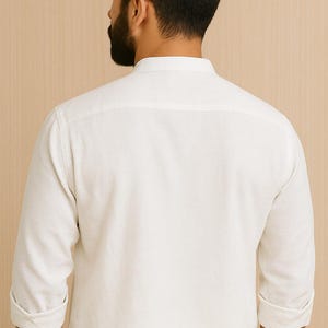 Pu&ograve; includere: Una camicia bianca a maniche lunghe con colletto alla coreana. Le maniche sono arrotolate all'altezza dei gomiti. La camicia &egrave; realizzata in un tessuto morbido e di colore chiaro. La schiena della camicia &egrave; visibile.