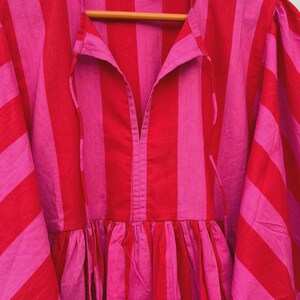 May include: A pink and red striped dress. The dress has a V-neck, gathered waist, and wide sleeves. The fabric appears to be lightweight. The dress is hanging on a wooden hanger.