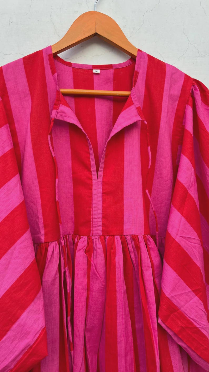 May include: A pink and red striped dress. The dress has a V-neck, gathered waist, and wide sleeves. The fabric appears to be lightweight. The dress is hanging on a wooden hanger.