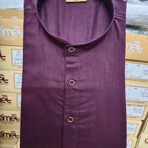 May include: A folded, dark burgundy shirt with a mandarin collar and three decorative buttons. The shirt has a tag that reads "comet 38". The shirt is part of a collection of ethnic wear.