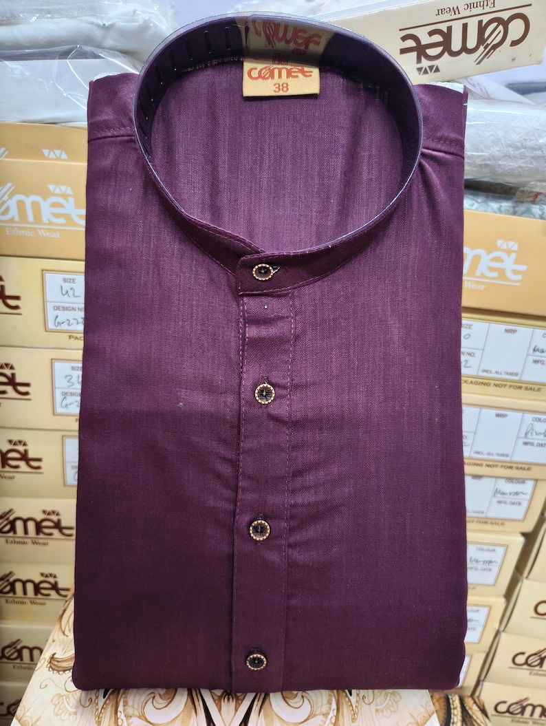 May include: A folded, dark burgundy shirt with a mandarin collar and three decorative buttons. The shirt has a tag that reads "comet 38". The shirt is part of a collection of ethnic wear.