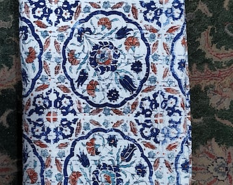 May include: Folded white textile with a repeating pattern of blue and orange floral designs. The pattern includes stylized flowers and leaves within ornate, curved frames. The textile is folded, showing the detailed stitching and the edges of the fabric.