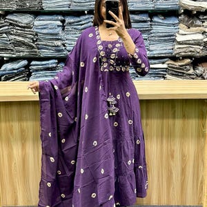 May include: A deep purple, two-piece outfit with a matching scarf. The dress features a square neckline, three-quarter sleeves, and a tiered skirt. The fabric is adorned with a white, circular pattern. The outfit is accessorized with a beaded necklace and tassel.