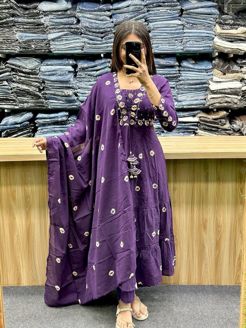 May include: A deep purple, two-piece outfit with a matching scarf. The dress features a square neckline, three-quarter sleeves, and a tiered skirt. The fabric is adorned with a white, circular pattern. The outfit is accessorized with a beaded necklace and tassel.