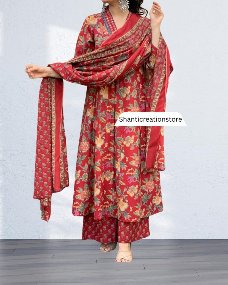 May include: A red floral print salwar kameez set. The set includes a long tunic top, wide-leg pants, and a matching scarf. The tunic has long sleeves and a V-neckline. The floral pattern features yellow, green, and pink accents. The scarf is draped over the shoulders. The text 'Shanticreationstore' is visible.